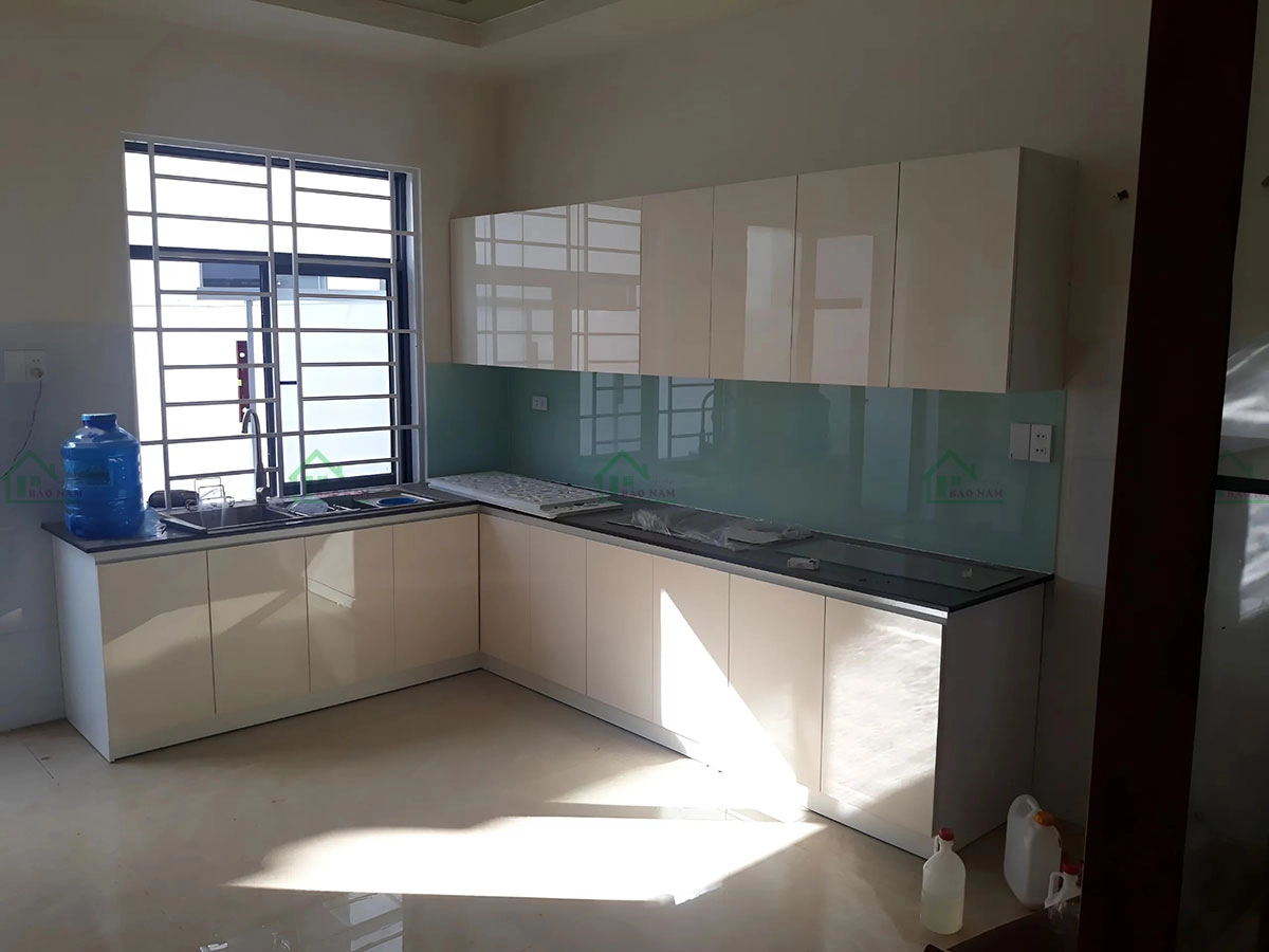 Tủ bếp acrylic bóng gương màu kem sáng, kết hợp bàn đá tối màu và kính ốp tường tạo điểm nhấn